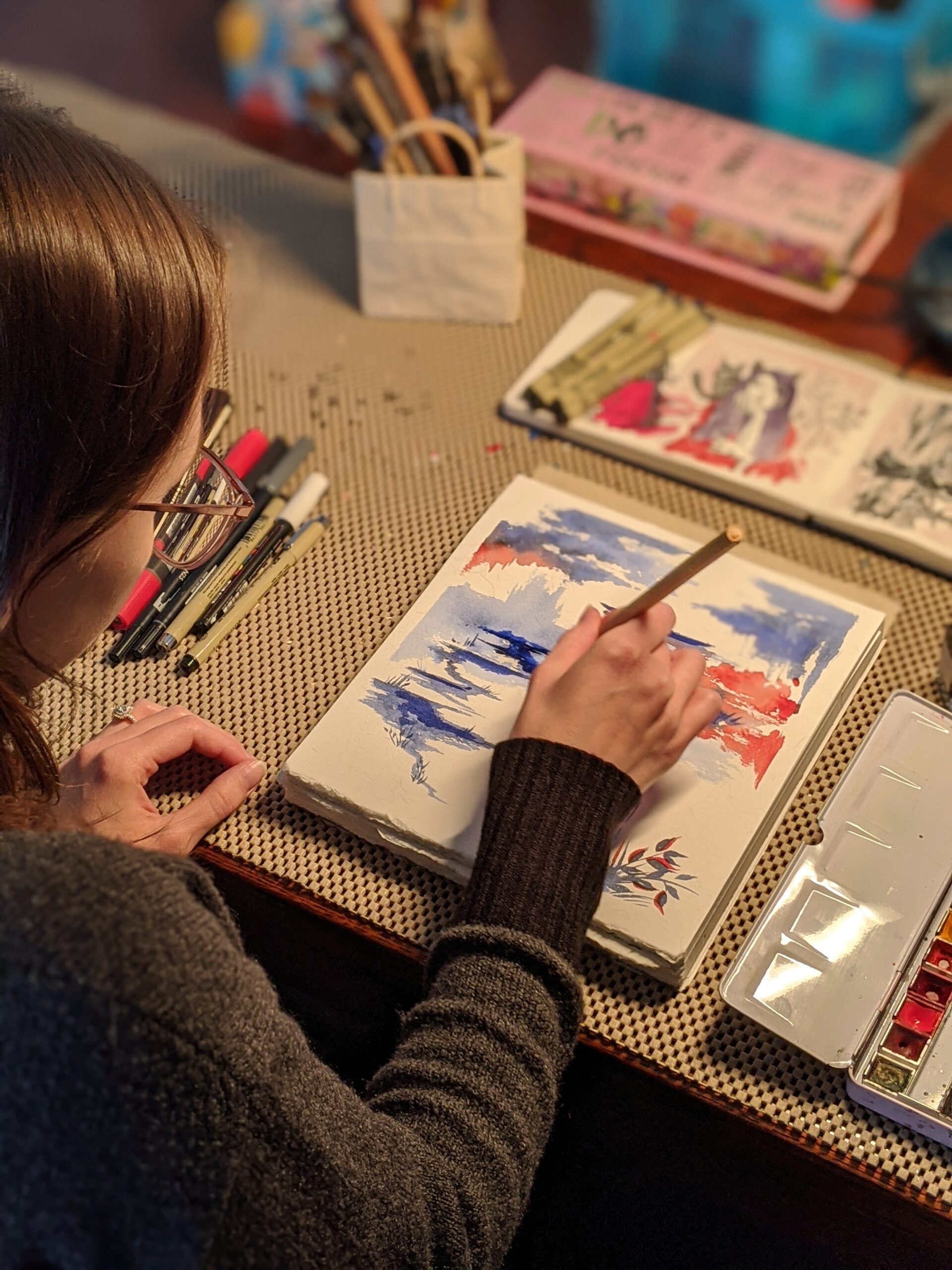 Artist painting with watercolors on paper in her studio on a wooden desk with a paintbrush in hand.